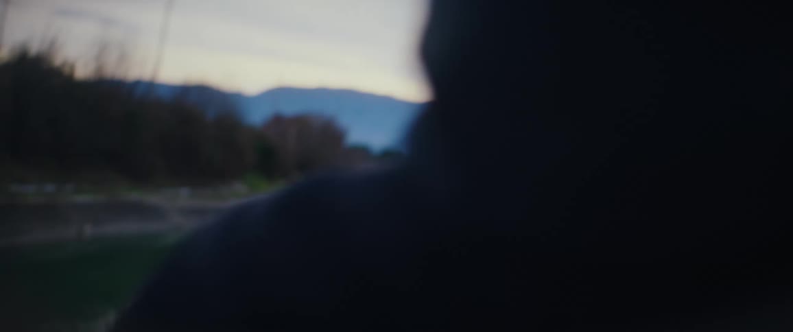 A young woman with long red hair smiles and looks back at a companion while walking beside a reflective river at dusk