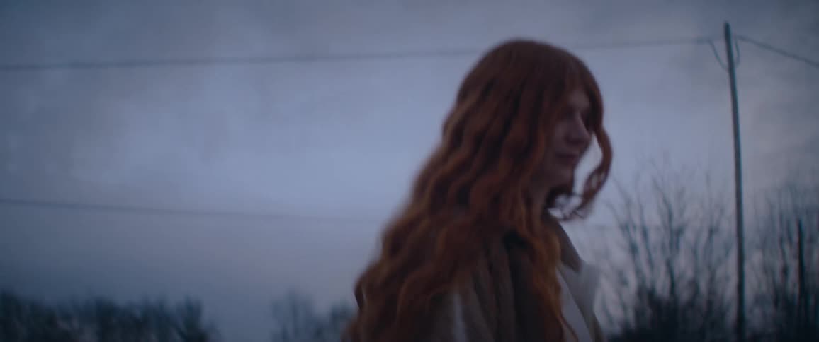 A view from behind shows a woman with long wavy red hair wearing a beige fleece jacket as she walks outdoors during twilight