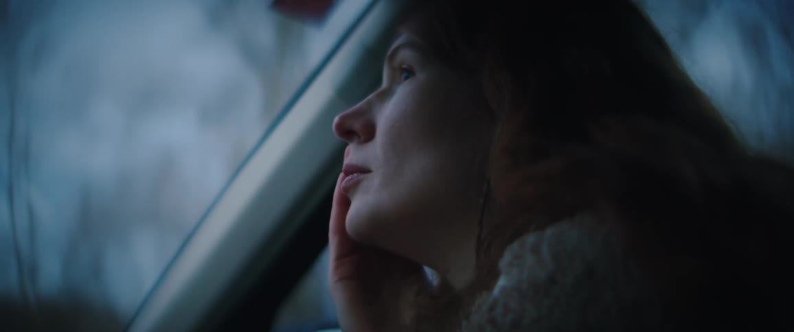 A close up shows a woman with long reddish brown hair wearing a beige fleece jacket as she looks out of a car window during twilight
