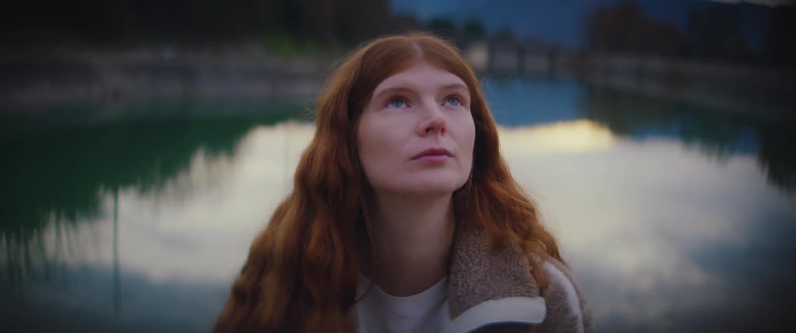 A woman with long wavy ginger hair wearing a fleece jacket looks up toward the sky with a thoughtful expression while sitting in front of a calm river reflecting the soft light of a blue twilight