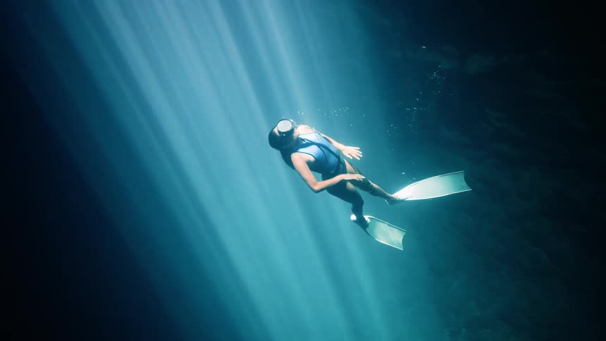 Female diver swimming underwater floats in water lit by rays of sunlight