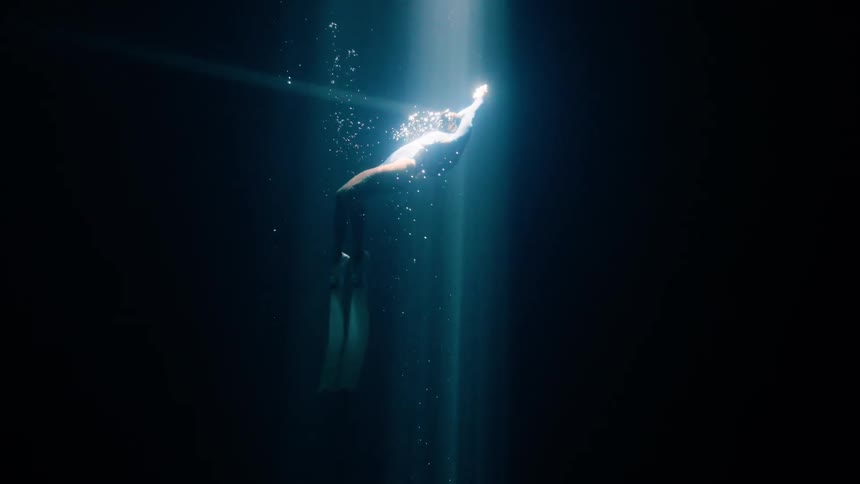 A person wearing long fins and a dark swimsuit swims upward through a bright beam of sunlight in deep dark water while small bubbles rise around them