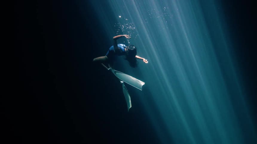 A female freediver floats gracefully in a dark underwater cavern while bright sun beams pierce through the surface to illuminate her body and long fins as she moves through the deep blue water with tiny bubbles rising around her head