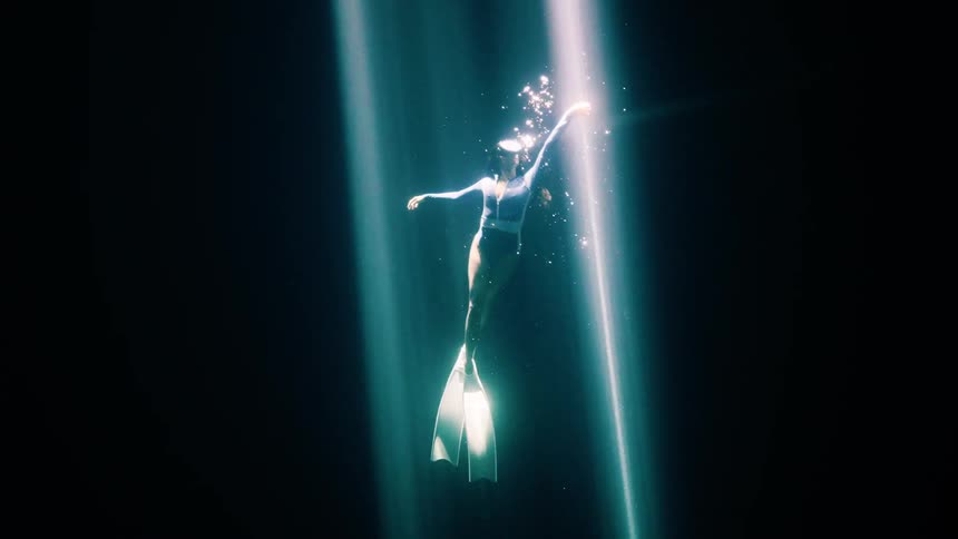 A free diver drifts weightlessly through dark water reaching into a beam of light as bubbles rise and the depths fall away into silence