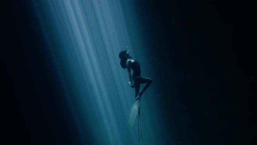 A lone free diver floats upward through deep blue water illuminated by soft beams of light that pierce the darkness like an underwater cathedral