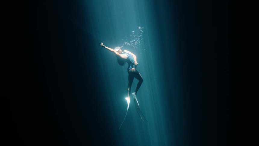 A diver with long fins ascends through a dramatic beam of light toward the waters surface