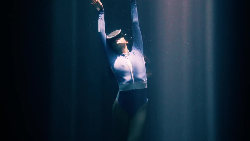 A woman wearing a blue swimsuit and diving mask drifts through a dark underwater while bright light beams shine down from the surface to illuminate her body and the small bubbles floating in the deep water around her outstretched arms