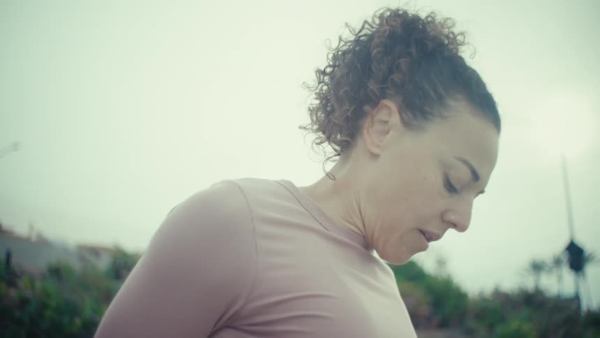 A young woman stretching her thigh by the handrail on a bridge at midday wearing a pink shirt