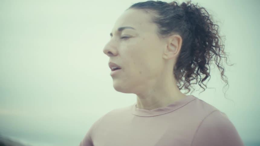 A woman with curly hair tied back wears a pink long sleeve top and rests her hand on a blue railing while looking out toward a misty ocean coastline after completing an outdoor morning workout on a cloudy day