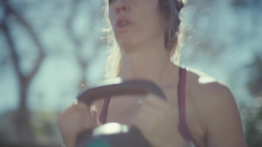 A close view shows the face of a woman with her hair pulled back as she performs squats while holding a kettlebell in a sunny outdoor area