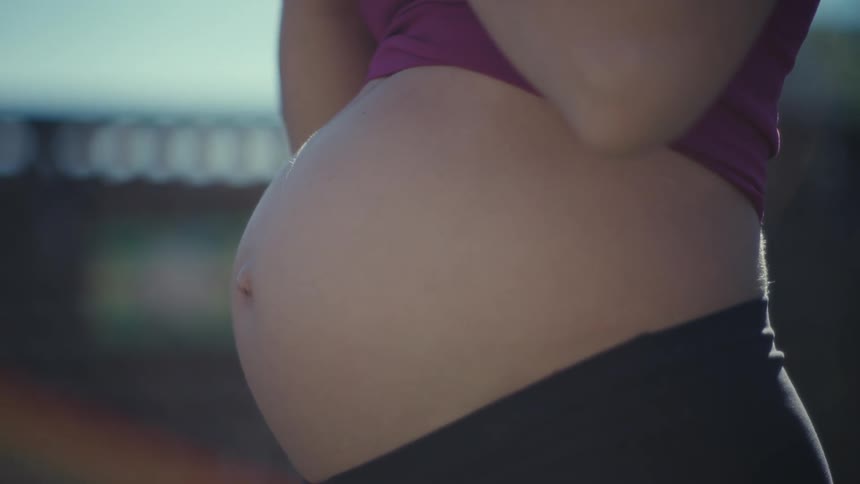 A young pregnant woman exercising outdoors doing squats while holding a dumbbell with her baby bump visible