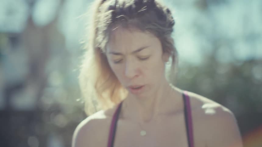 A close up shot features a pregnant woman during an intense outdoor workout and she breathes heavily with a focused expression while sunlight flares around her blonde hair and she stands in a backyard setting wearing a pink top and black leggings