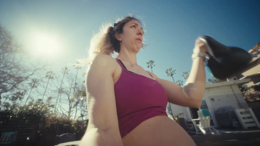 A pregnant woman lifts a kettlebell in bright morning sunlight her focused expression and strong posture framed against a clear blue sky