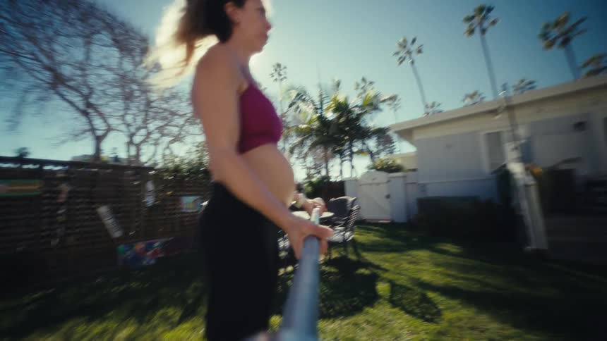 A view from a low angle shows a pregnant woman with her hair in a ponytail as she picks up a barbell and performs several repetitions of an exercise in a sunny backyard