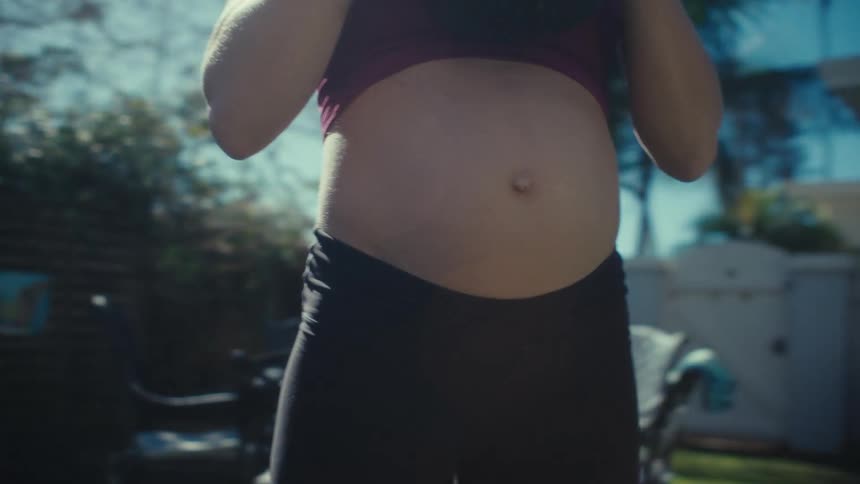 A close view shows the bare belly of a pregnant woman wearing a red sports bra and black leggings as she performs squats while holding a kettlebell in a sunny outdoor area