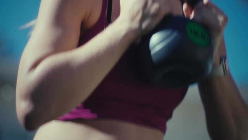 A close up shot captures a pregnant woman during a workout session and she holds a kettlebell near her chest while breathing deeply with a determined expression on her face under a clear blue sky and bright sunlight in an outdoor setting