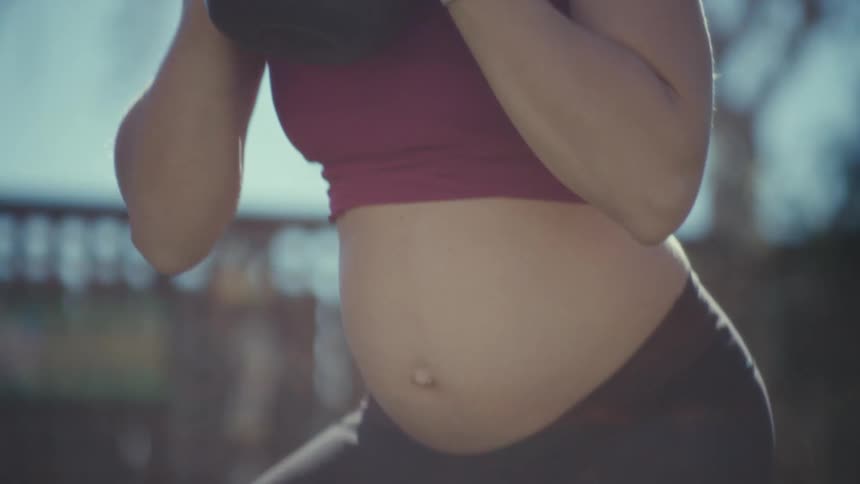 A pregnant woman performs kettlebell goblet squats outdoors in a sunlit settingthe camera focuses closely on her form and breathing as she moves through the repetitions