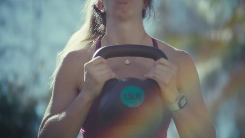 Adult pregnant woman holding a kettle bell while squatting with trees on the background outdoros during day time