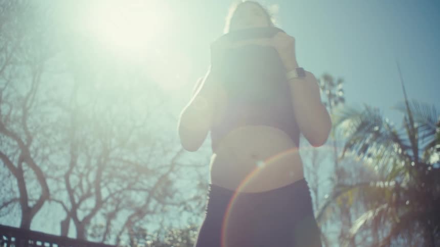 A view from a low angle shows a pregnant woman with her hair in a ponytail as she picks up a kettlebell and performs several repetitions of an exercise in a sunny backyard