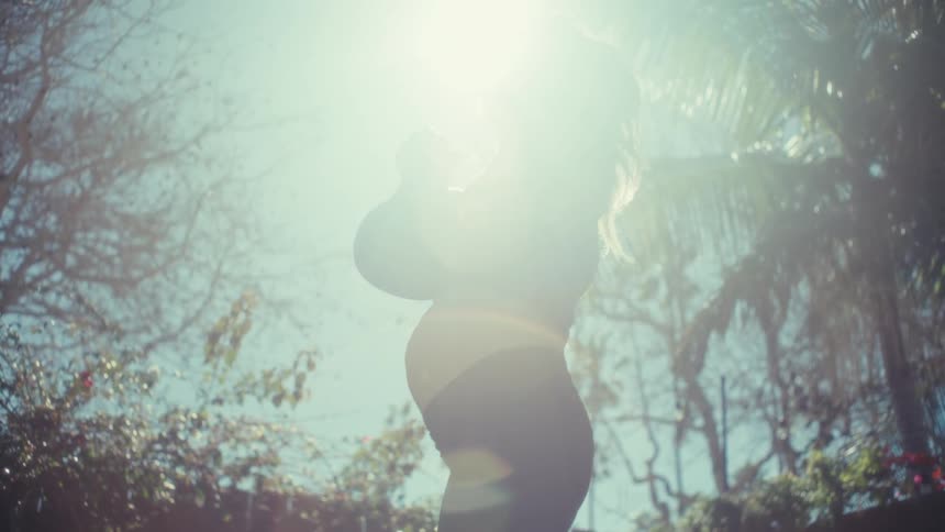 A pregnant woman performs kettlebell squats outdoors in the bright flared sunlight