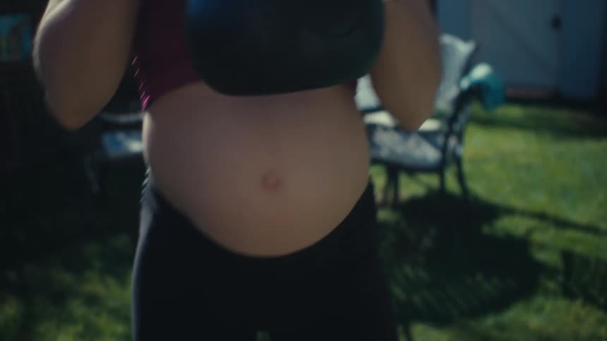 A pregnant woman does squats in her yard while holding a kettle bell weight