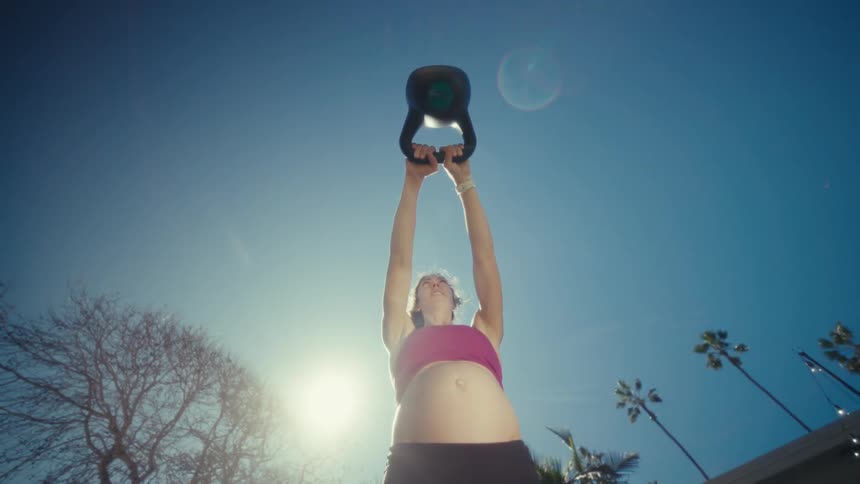 Adult pregnant woman lifting a kettle bell in a lawn outdoors during day time