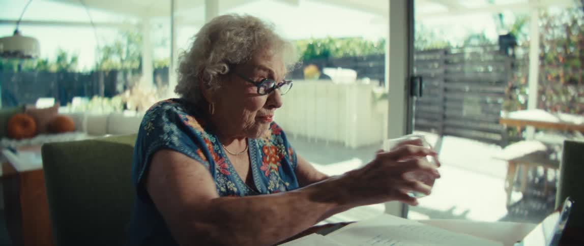 An elderly woman drinks water and performs a seated stretch in a bright modern home