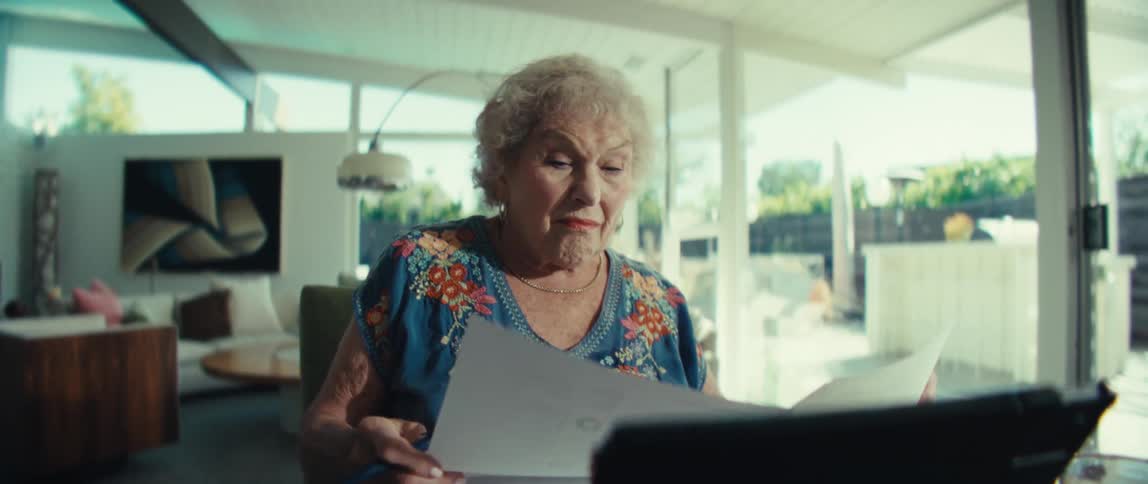 Elderly woman sitting down reading a piece of paper in front of a table with a tablet computer and a calculator in a living room outdoors during day time