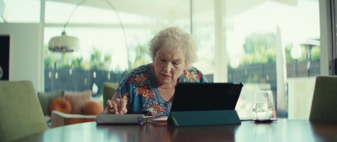Elderly adult woman using a calculator and a tablet computer over a wooden table in a living room indoors during day time