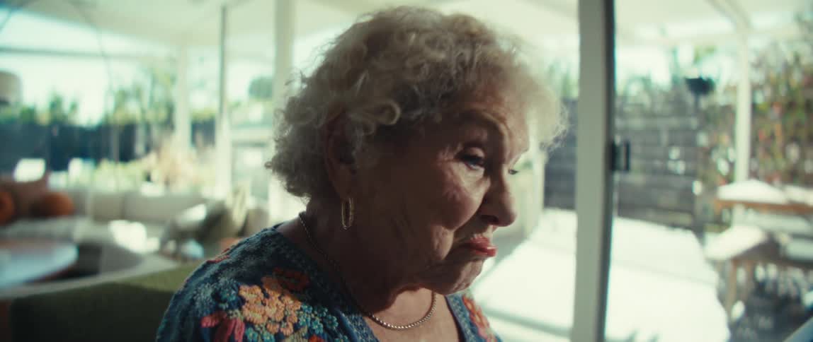 An older woman with curly gray hair wearing a blue floral shirt sits at a wooden table and looks at a paper document with a serious expression inside a bright room with large windows