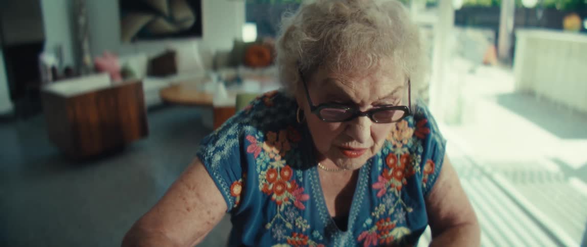 An elderly woman wearing a blue floral blouse sits at a wooden table reviewing financial documents and bar charts using a tablet