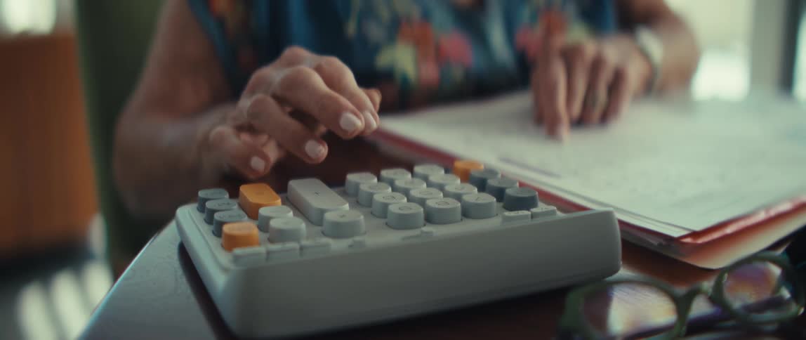 An elderly woman types on a chunky gray calculator while looking at paperwork on a wooden desk beside an open notebook and reading glasses
