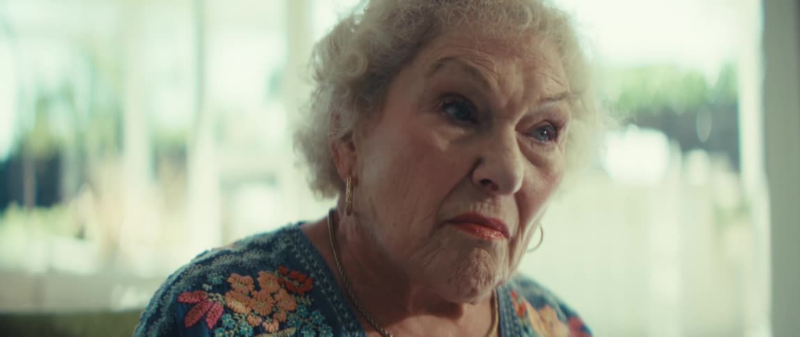 An elderly woman sitting indoors reading a document she is holding with a serious expression at midday