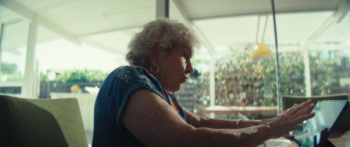 An older woman with curly white hair wearing a blue floral shirt sits at a table and looks toward a tablet while speaking