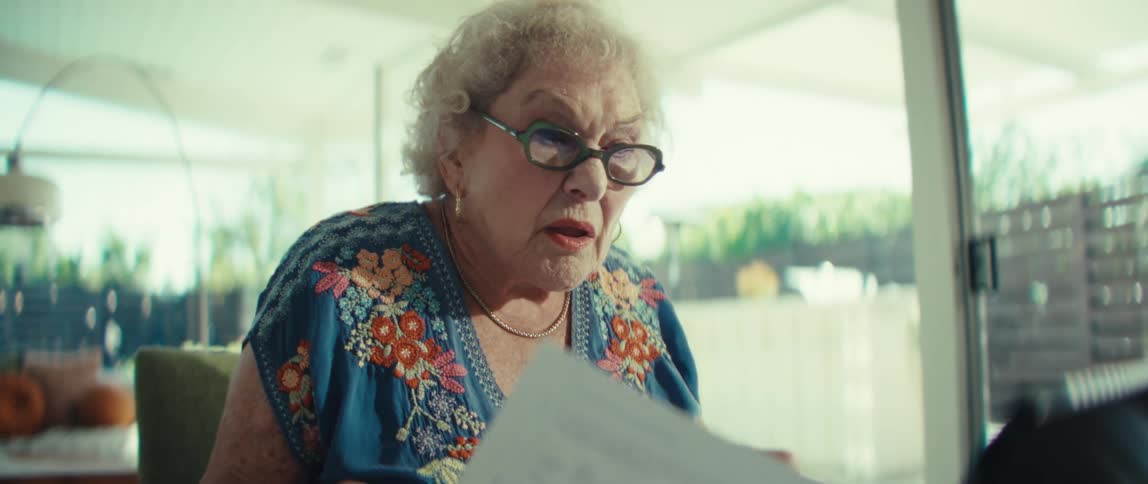 An elderly woman with white hair and glasses looks intently at a document she is holding and talks on a video call