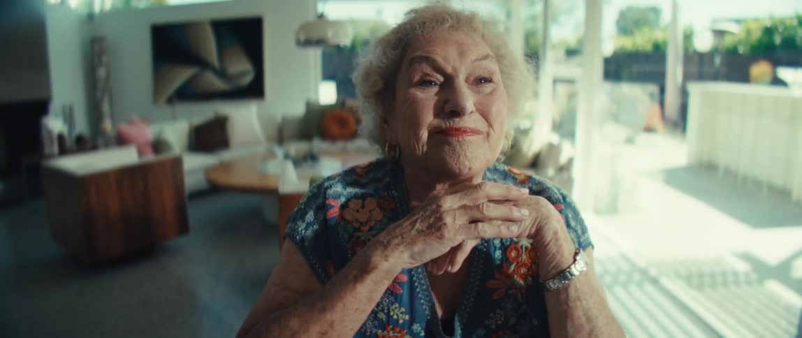 An elderly woman takes off her glasses and smiles warmly at the camera while resting her chin on her hands