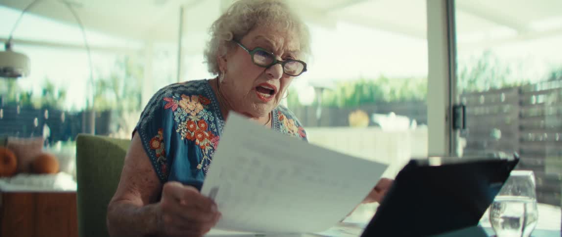 An older woman wearing glasses and a floral shirt sits at a table and looks at a document while speaking in front of a tablet