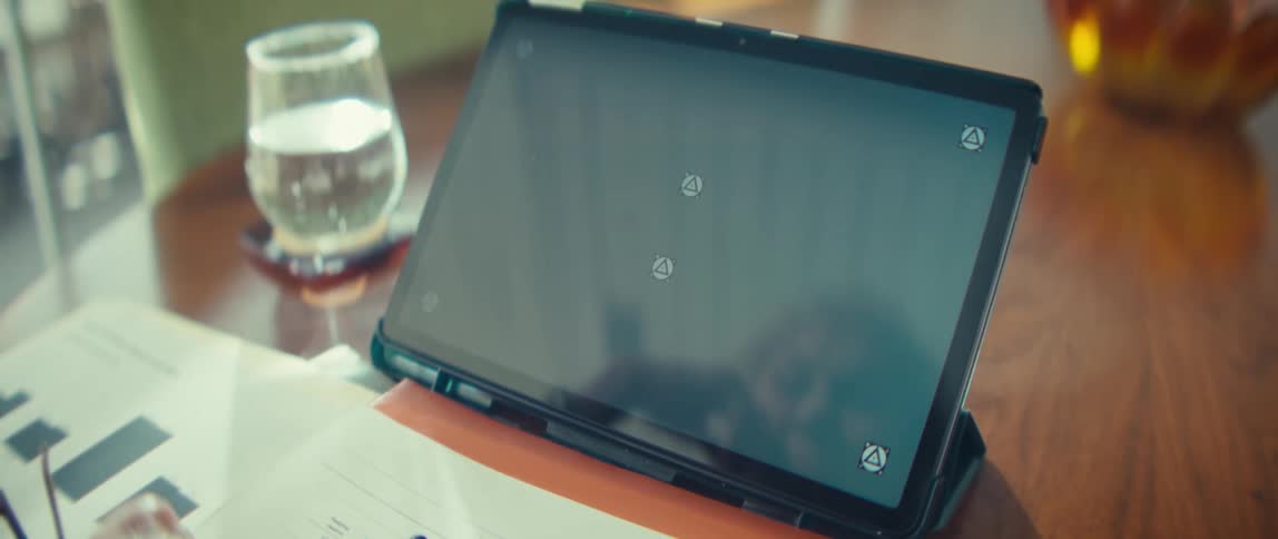 A close view shows a tablet with a black screen and several small tracking markers on it sitting on a wooden table next to paper documents and a glass of water