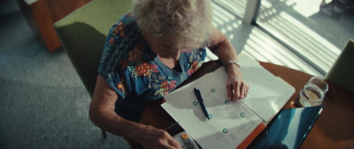 A view from above shows an older woman with curly white hair wearing a blue floral shirt as she uses a calculator and reviews financial documents while sitting at a table with a tablet and a glass of water