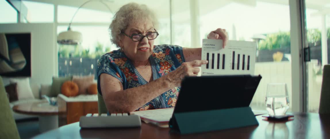 Elderly woman references a printout of charts while using a tablet as she sits at a table at home