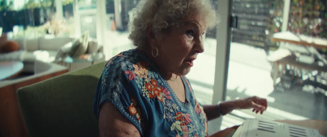 An elderly woman with curly hair wearing a blue floral blouse sits at a wooden table reviewing charts and documents then smiles broadly
