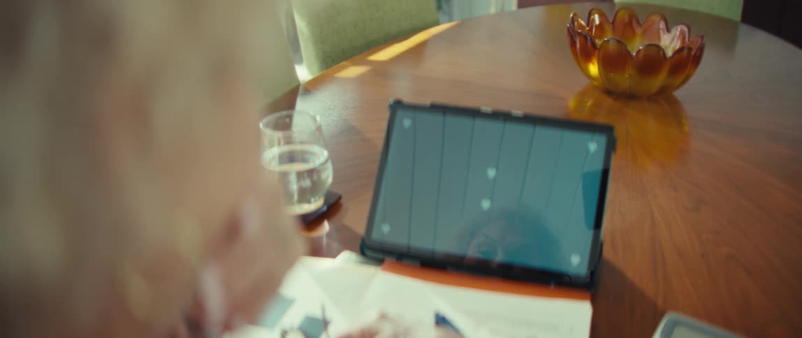 Woman sits at a table in front of a tablet computer with paperwork and a glass of water