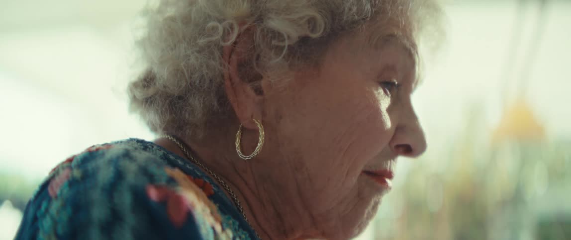 An elderly woman with curly white hair and gold hoop earrings is shown looking over some documents the camera focuses on her hands as she holds and examines a sheet titled tradeoff analysis which features various charts and graphs