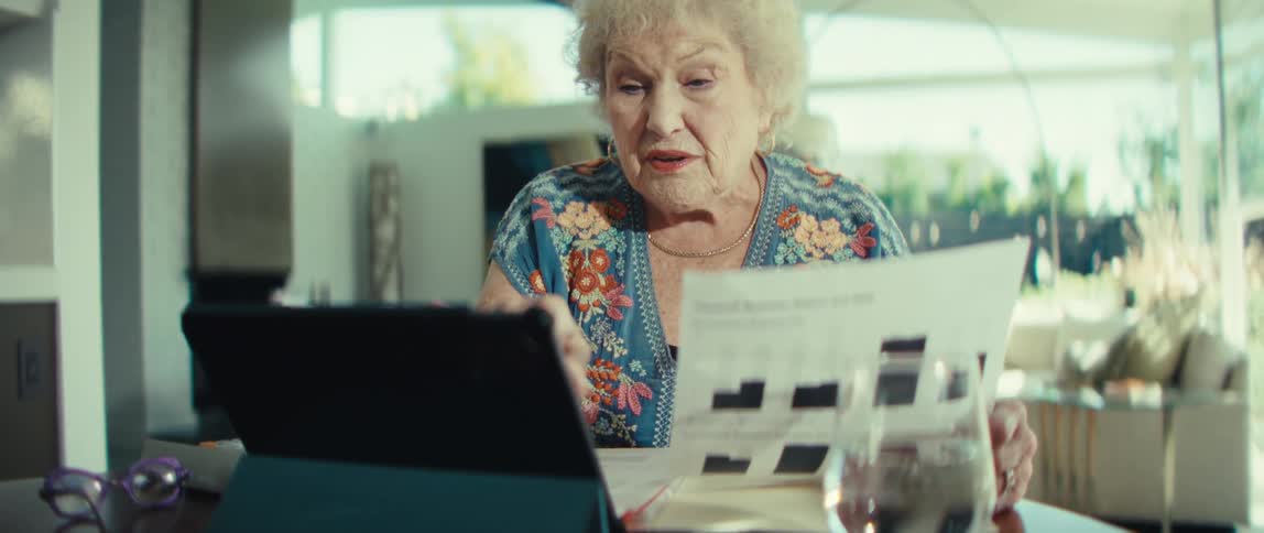 An elderly woman sits at a table and holds a financial document toward a tablet screen during a video call while explaining the charts on the paper