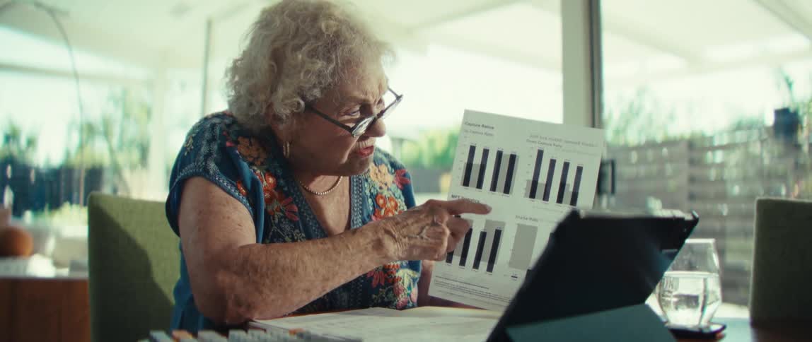 An elderly woman with glasses studies a printed page of bar charts holding it up thoughtfully beside a tablet as sunlight fills the quiet room