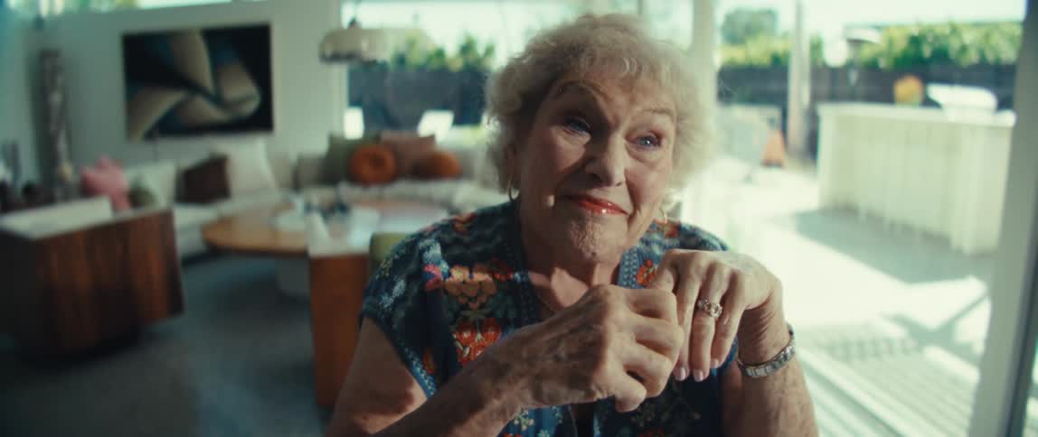 An older woman with curly white hair wearing a floral blue shirt sits indoors with her hands clasped while looking ahead with a soft and thoughtful expression