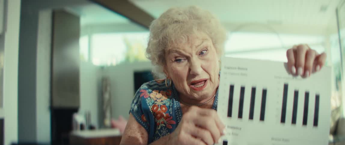 An elderly woman with curly white hair wears a blue floral blouse while holding a financial document during a video call on her tablet
