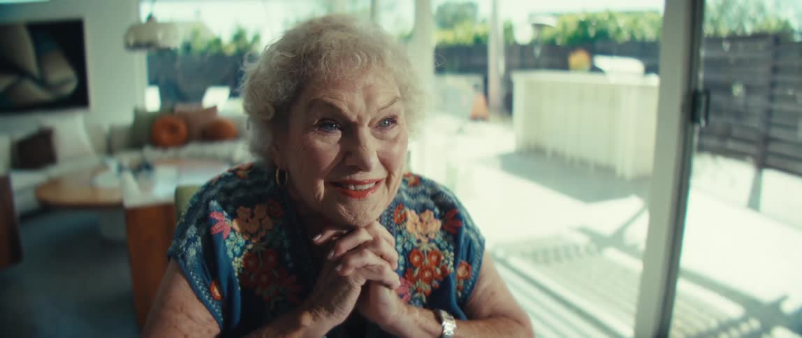 An elderly woman with curly white hair sits in a bright living room and she takes off her glasses and smiles warmly while clasping her hands together