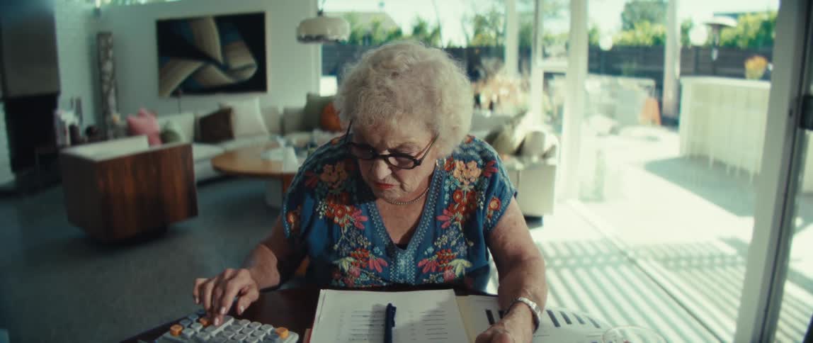 An elderly woman focuses on her finances using a calculator and tablet in a sunlit modern living room