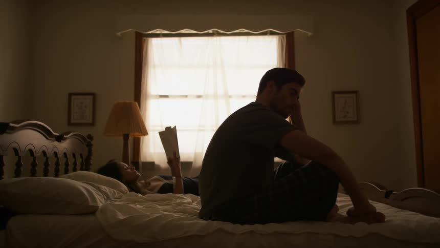 A woman reads a book in bed while a man sits beside her
